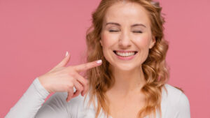 Mulher loira sorrindo e apontando para a bochecha em fundo rosa, representando alívio após extração de dente do siso na Ferdinand Odontologia.