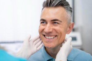Homem sorrindo sendo examinado por mãos com luvas odontológicas. Veja quais produtos usar para limpar a prótese protocolo em Bauru.